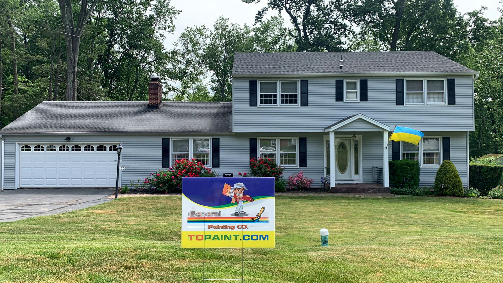 Blue-gray colonial home with vibrant flowers and yard sign